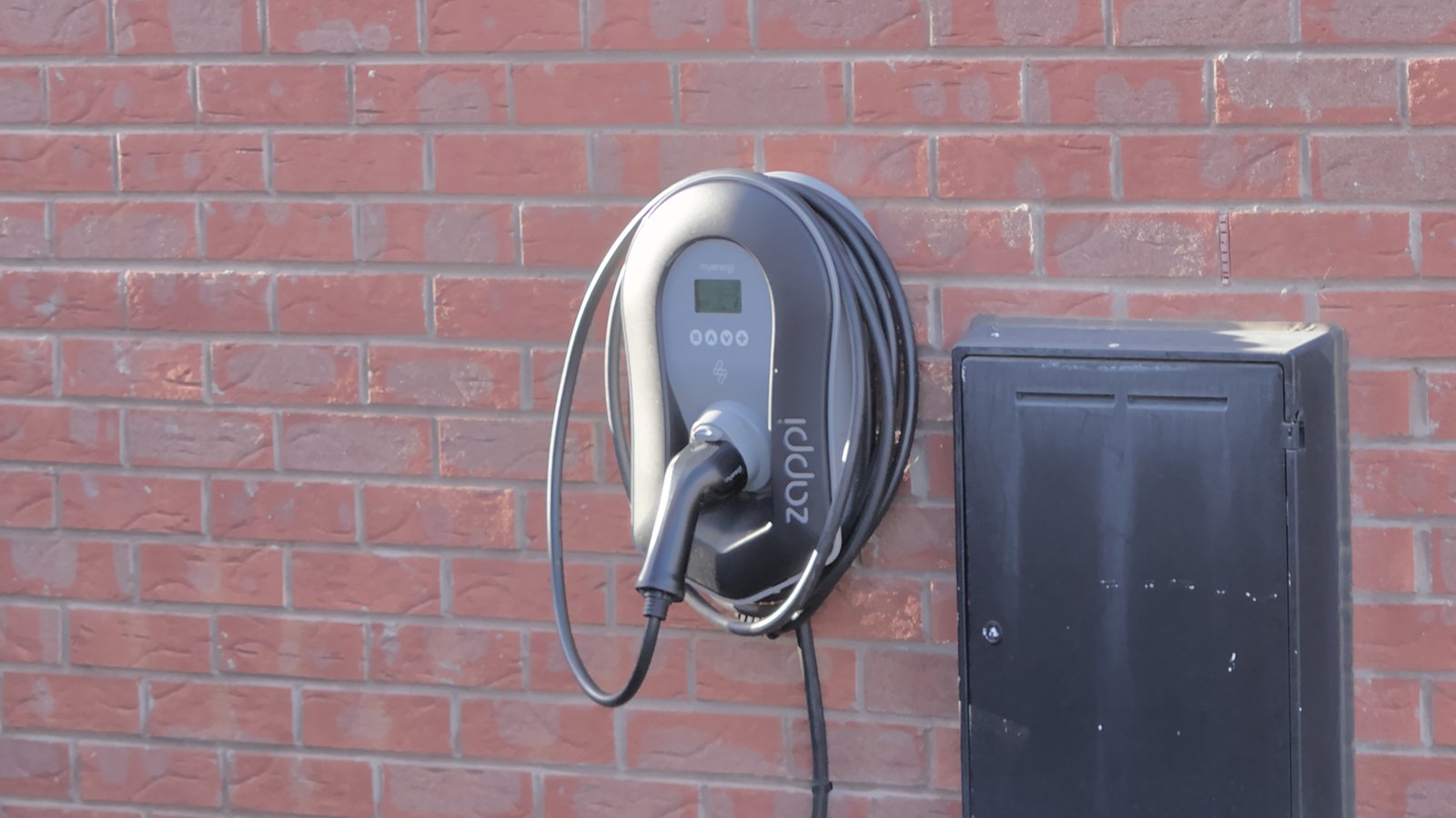 A wall-mounted home electric car charger with a tethered cable on an outside brick wall